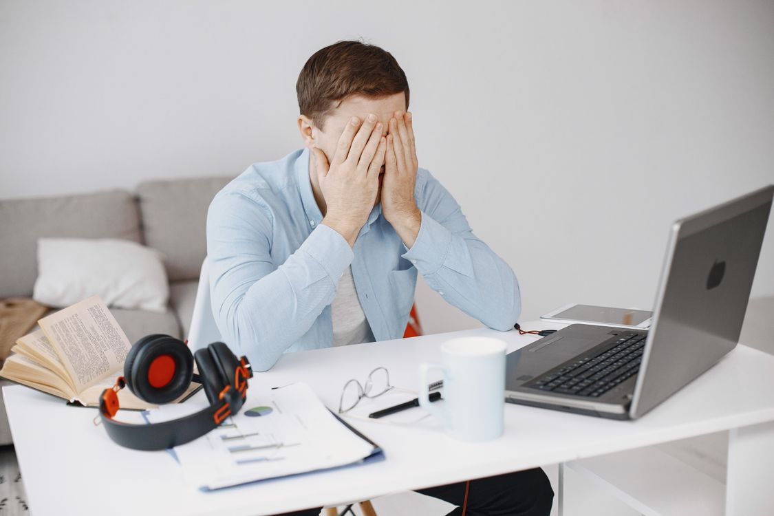 A frustrated man sits at his home office desk holding his head while looking at his laptop screen.